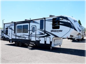 White and navy Momentum travel trailer with large slide-out room displayed at RV dealership