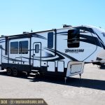 Front angle view of white and navy Momentum M-Class toy hauler with open garage door and slide-out cargo area at RV sales lot