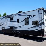 White and navy Momentum M-Class travel trailer with dual axles parked at Royal Coach Sales lot, featuring sleek curved graphics and modern design