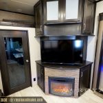 RV living area with flat-screen TV mounted above fireplace insert, dark wood cabinetry, and bedroom doorway visible