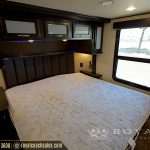 Luxury RV master bedroom with queen bed, dark wood headboard with blue accent lighting, large window, and overhead cabinets