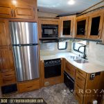 Full RV kitchen layout showing stainless steel refrigerator, black stovetop, microwave, wooden cabinets, beige countertops, and multiple windows
