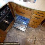 Open dishwasher drawer with blue racks in RV kitchen next to black stovetop and beige countertops with wooden cabinetry