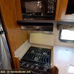 RV kitchen interior with stainless steel refrigerator, black stovetop with cookware rack, beige countertops, wooden cabinetry, and microwave mounted above