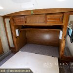 RV bedroom interior with wooden cabinetry, bed with white mattress, gray carpet flooring, and storage cabinets above