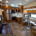 Interior view of luxury RV showing living area with brown leather seating, wood cabinetry, kitchen with stainless steel appliances, and dining booth
