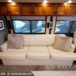 Luxury RV lounge with cream leather sofa, decorative pillows, large windows with shade covers, and polished wooden cabinetry