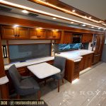 Full RV interior view showing dinette seating area, wood cabinetry throughout, kitchen counter, large windows, and wooden beam ceiling with LED lighting