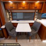 RV dining area with wood cabinetry, two gray upholstered chairs flanking a white table, and large window with retractable shade