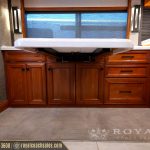 Luxury RV kitchen with rich wood cabinetry, white countertop sink, and large window with blue mesh shades in a modern motorhome interior