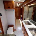 RV bathroom and hallway featuring toilet, dual stainless steel sinks, wood vanity, shower enclosure, and beige tile flooring