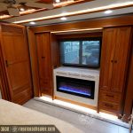 Wide-angle view of luxury RV living area showing entertainment center with TV, electric fireplace, and wooden built-in storage cabinets on both sides