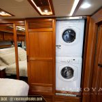 RV laundry area with stacked washer and dryer units, wooden cabinetry, and mirrored closet door with queen bed visible
